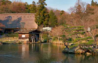 A serene Japanese garden features a traditional wooden watermill by a calm pond. The scene is framed by lush green trees and a thatched-roof building in the background. The soft sunlight enhances the tranquil atmosphere.