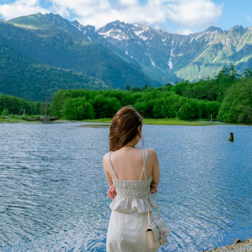 Kamikochi, Japan A woman in a white dress stands by a serene lake, facing away from the camera. She is surrounded by lush green trees, and majestic snow-capped mountains are visible in the background under a partly cloudy sky. The scene is peaceful and picturesque.