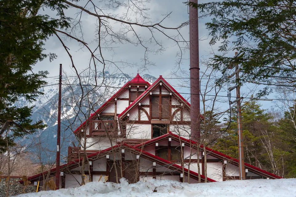 A traditional wooden house with a red roof and white walls stands amidst leafless trees and snow. A towering mountain is visible in the background under a cloudy sky, creating a picturesque winter scene. A tall chimney-like structure is seen on the right side of the house.