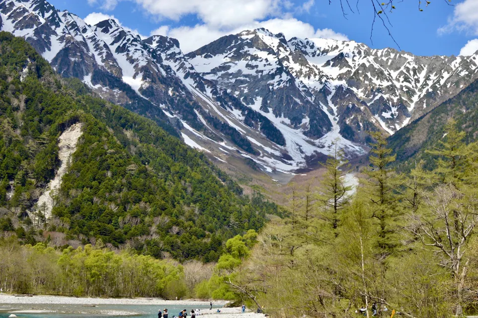 A beautiful landscape featuring a majestic mountain range with snow-covered peaks under a blue sky. A lush green forest stretches at the base of the mountains, with a river flowing through it. People can be seen walking by the riverbank, enjoying the view.