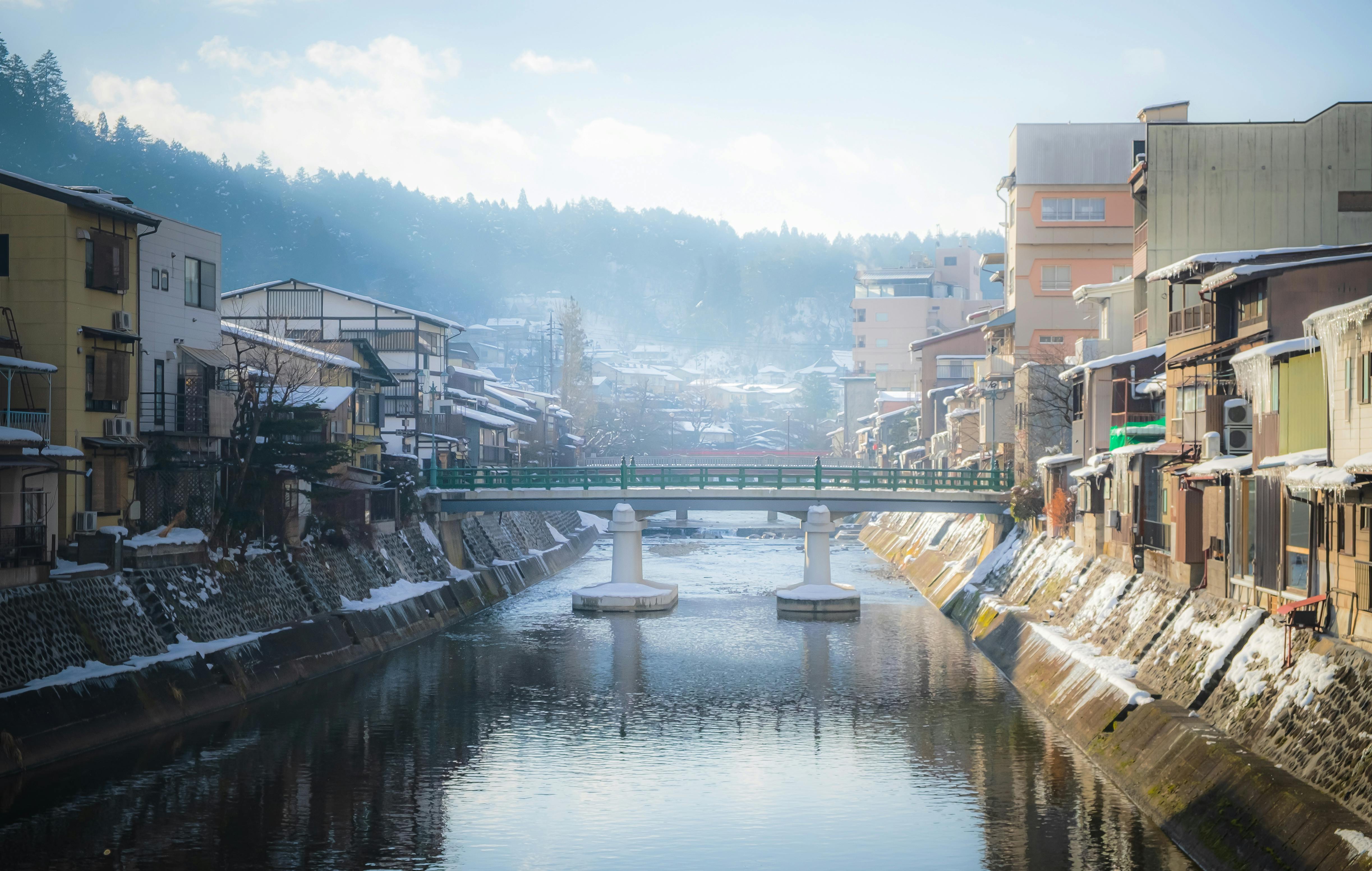 A quiet river flows through a Japanese town with traditional wooden houses and buildings lining both sides. A green bridge crosses the river, and light snow covers rooftops and sidewalks under a soft, misty sky.