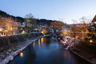 A river runs through a small town at dusk, lined with stone walls and softly illuminated streetlights. Leafless trees and houses sit on both sides, with snow lightly covering the ground.