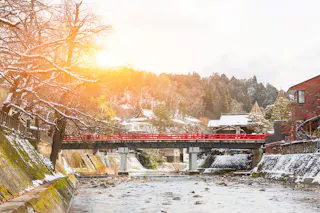 A sunlit scene of a red bridge crossing a shallow river, with snow-dusted trees and traditional Japanese buildings in the background.