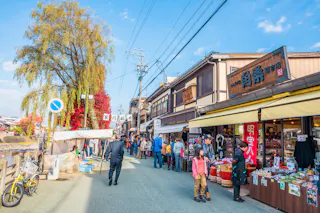 People walk along a lively street lined with shops and market stalls in a traditional Japanese town. Trees with autumn foliage and clear blue sky are visible, creating a vibrant, bustling scene.
