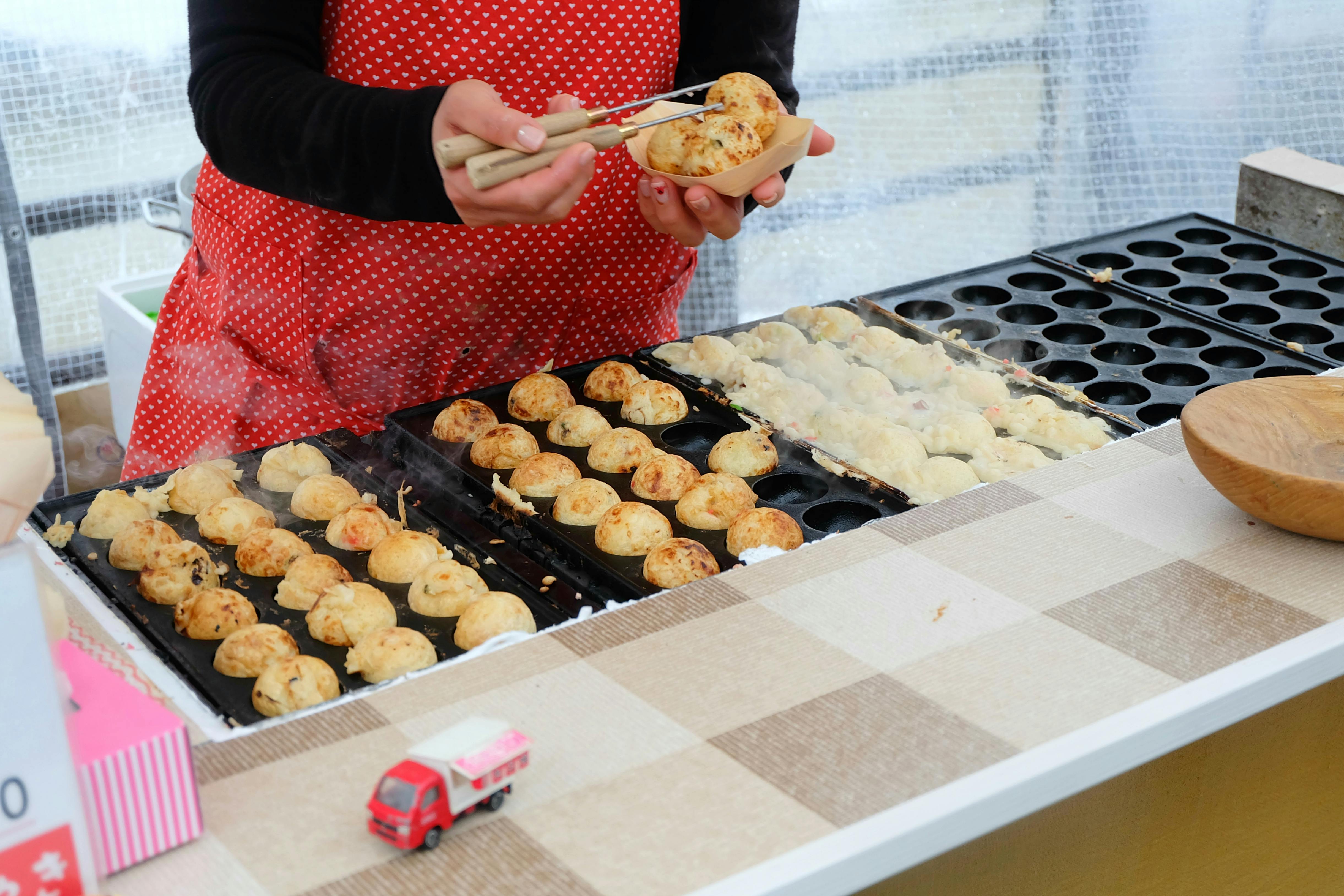 A person in a red polka dot apron uses tongs to prepare takoyaki balls on a hot griddle at a food stall. A toy food truck is on the counter nearby.
