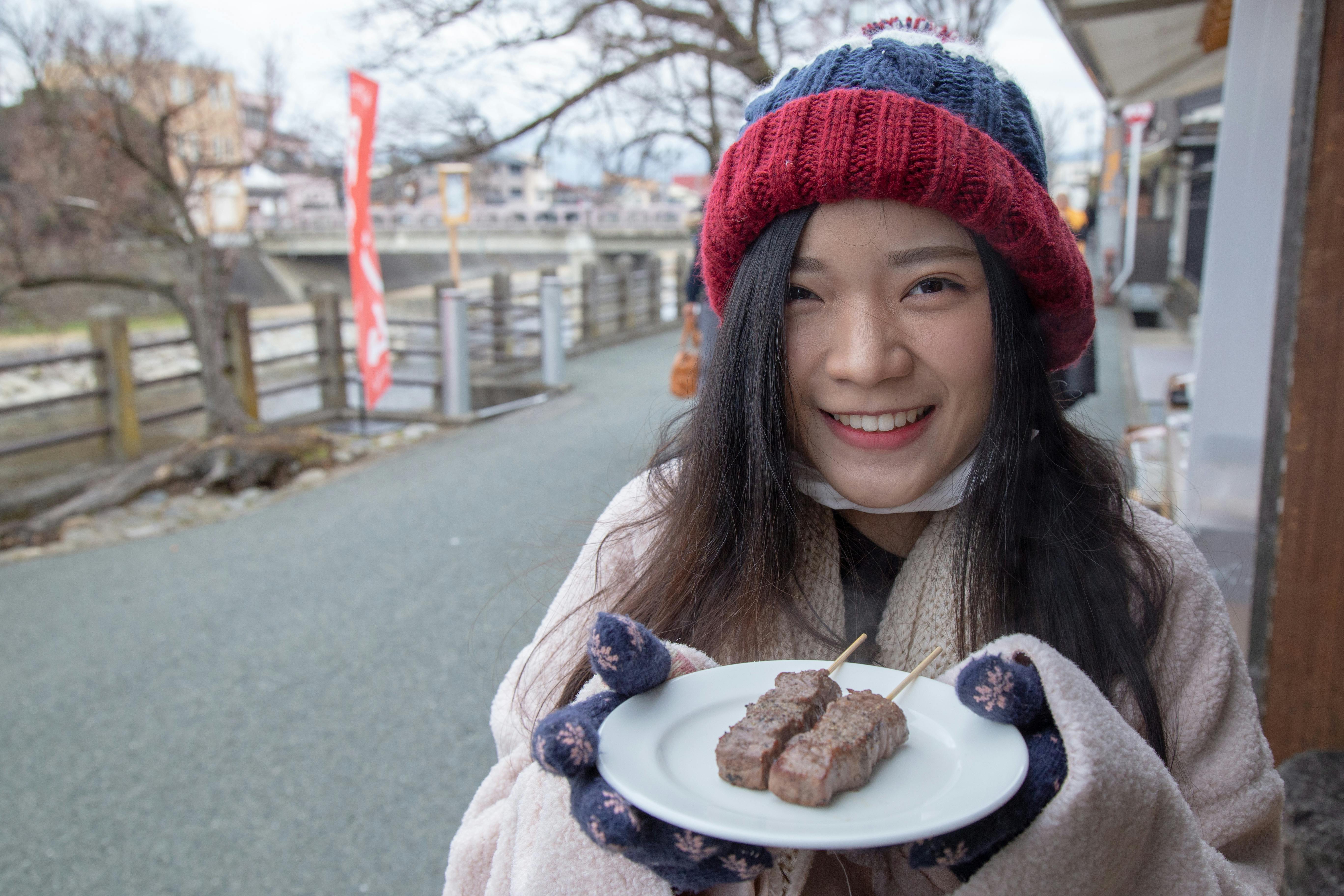 A person in a red and blue beanie and gloves smiles while holding a plate with skewered grilled meat. They stand on a walkway, with wooden railings and trees in the background, suggesting a cool outdoor setting.