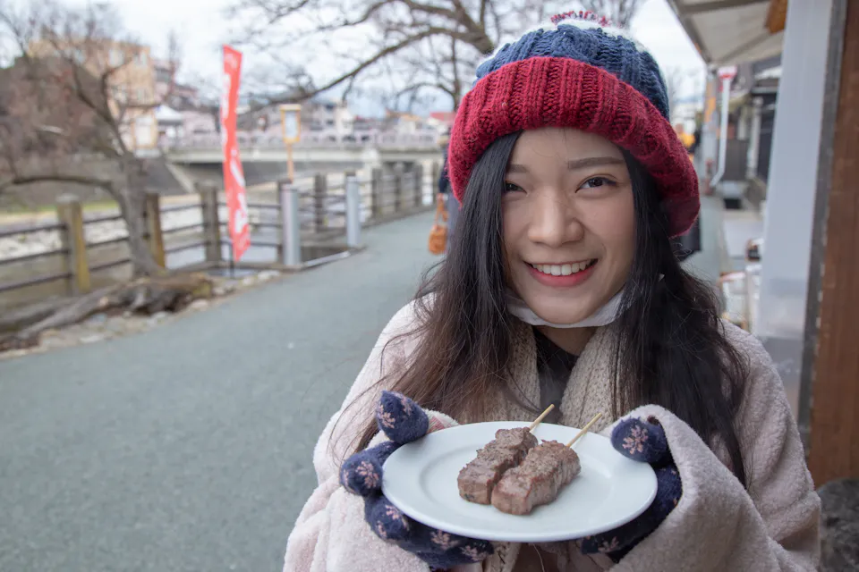 Miyagawa Morning Market A person in a red and blue beanie and gloves smiles while holding a plate with skewered grilled meat. They stand on a walkway, with wooden railings and trees in the background, suggesting a cool outdoor setting.