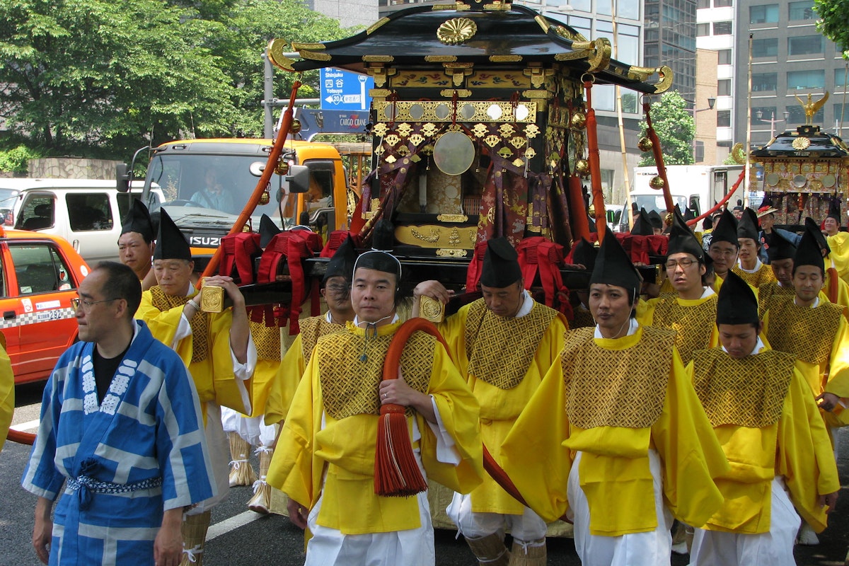 Spring Takayama Festival (Sanno Matsuri) A group of people dressed in traditional yellow and blue attire participate in a street parade, carrying an ornate, gold-adorned palanquin. Buildings and a tree-lined street form the backdrop, with several onlookers and vehicles visible.