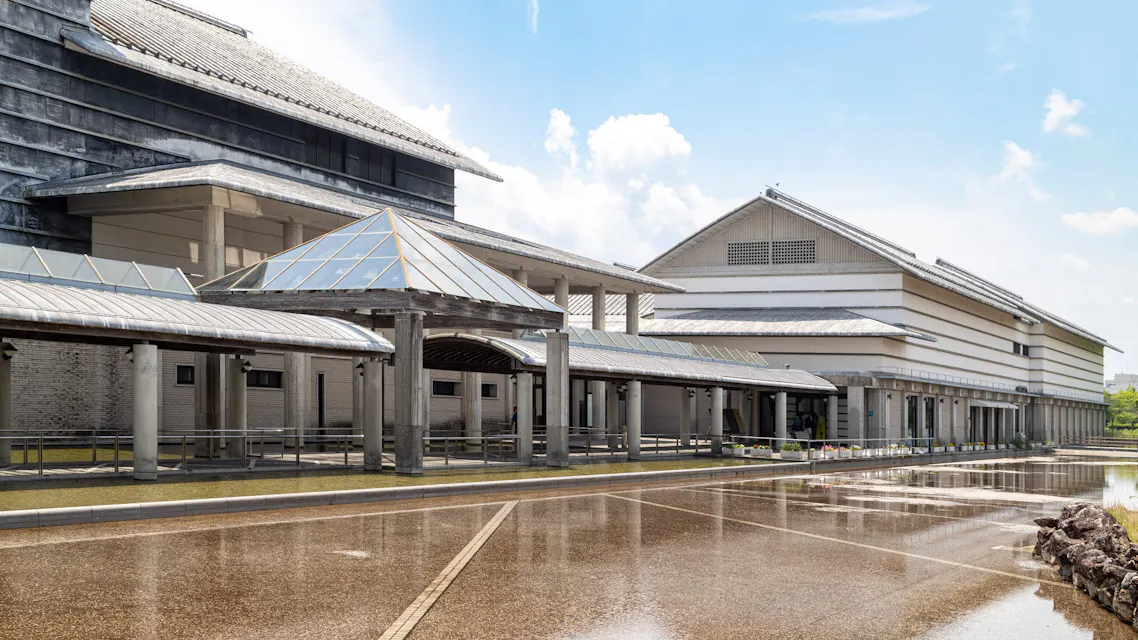 A modern architectural building with a traditional Japanese design features multiple structures connected by covered walkways. The exterior showcases clean lines, large windows, and a combination of concrete and tiled roofs. The foreground has a wet pavement.