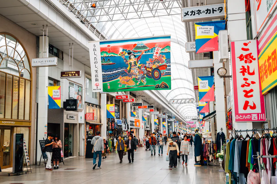 A bustling indoor shopping street in Japan, with colorful banners and signs in Japanese hanging from above. Shoppers walk along the tiled pathway, flanked by various stores and boutiques on both sides. A large, vibrant tapestry is prominently displayed overhead.