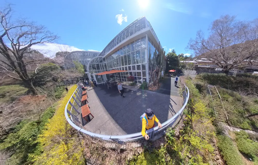 A spherical panorama image of a greenhouse building with large glass windows, taken on a sunny day. People are seen walking and sitting around the patio area outside. The surrounding landscape is lush with greenery, and trees with bare branches are visible.