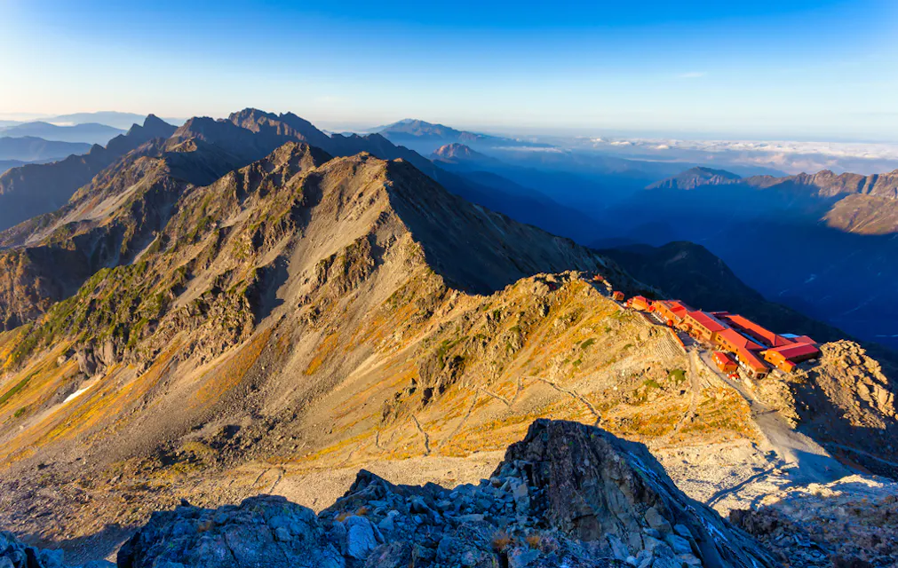Mount Yari A panoramic view of a rocky mountain range under a clear blue sky. A red-roofed structure stands on a ridge, nestled amid the rugged terrain. Sunlight casts vibrant hues across the landscape, illuminating the peaks and the surrounding valley.