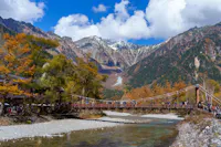 A scenic mountain landscape with a suspension bridge stretching over a clear river. The area is surrounded by colorful autumn trees and towering snow-capped peaks under a clear blue sky with scattered clouds. Many people are walking on the bridge.