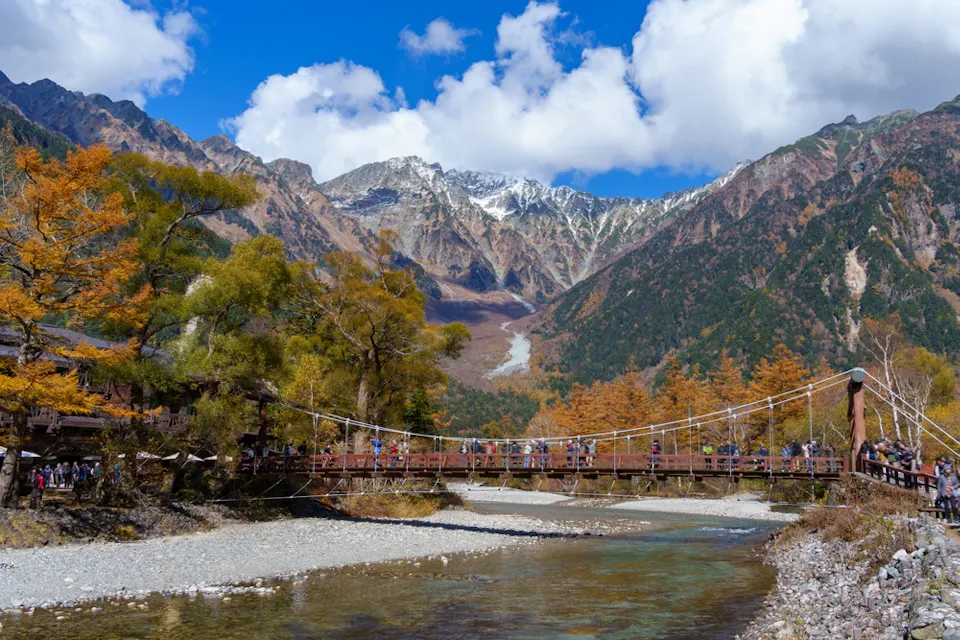 What Will I Find at Kamikochi A scenic mountain landscape with a suspension bridge stretching over a clear river. The area is surrounded by colorful autumn trees and towering snow-capped peaks under a clear blue sky with scattered clouds. Many people are walking on the bridge.