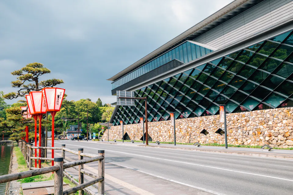Kochi Castle Museum of History A modern building with a geometric glass facade stands adjacent to a stone wall along a quiet street. Traditional red lanterns line the sidewalk, and a fence borders the roadside. Trees and greenery are visible in the background under a cloudy sky.
