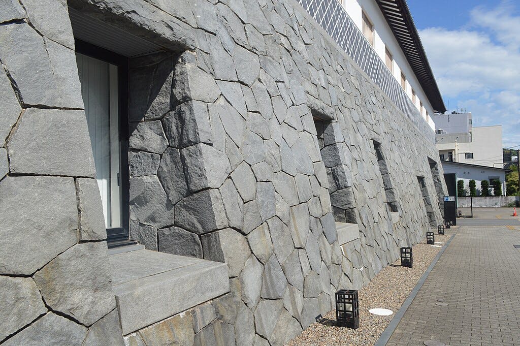 A stone wall with irregularly shaped gray stones, small rectangular windows, modern lanterns along a gravel path, and a cityscape with buildings in the background, under a blue sky with clouds.