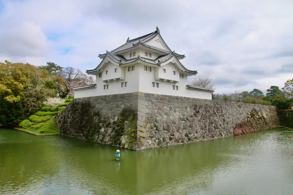A traditional Japanese castle stands on a stone foundation surrounded by a green moat. The white structure features intricate, curved eaves and an ornate roof. Trees and foliage are visible in the background under a partly cloudy sky.