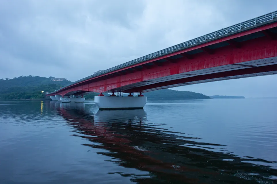 A large red bridge with white supports spans over calm water, reflecting its vivid color in the surface below. The sky is overcast, and the distant shoreline is lined with trees and hills. The scene is peaceful and serene.
