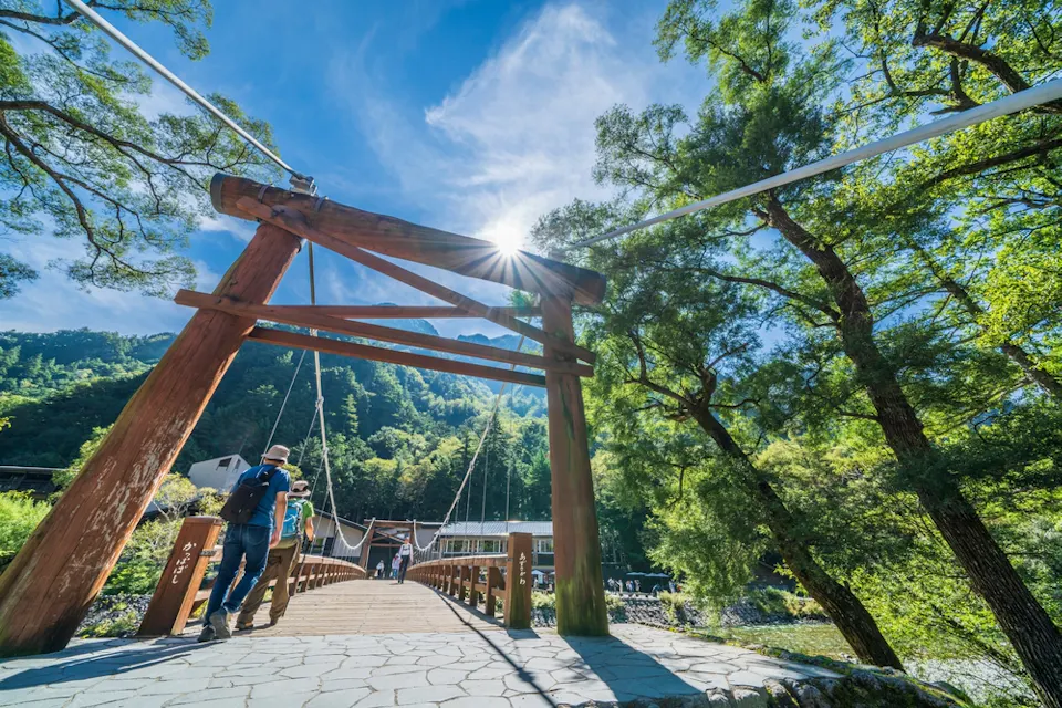 Kappa Bridge A person with a backpack crosses a wooden suspension bridge under a large traditional wooden torii gate. The bridge is surrounded by lush greenery, and the sun shines brightly from a blue sky, casting shadows on the stone path leading to the bridge.