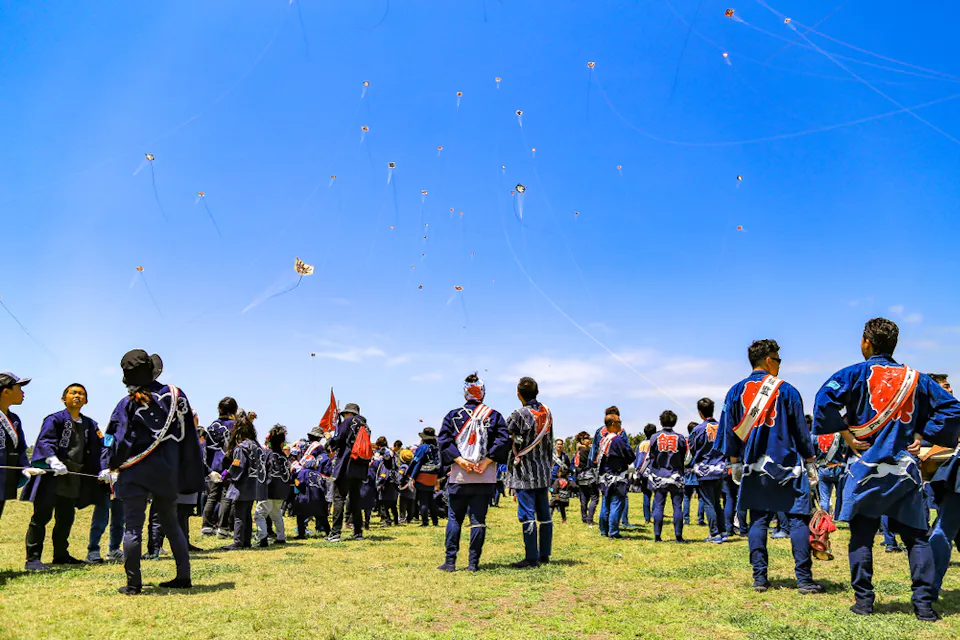 A group of people in traditional clothing are gathered in a grassy field, watching numerous kites flying in the blue sky. The vibrant scene suggests a festive or cultural event under bright daylight.