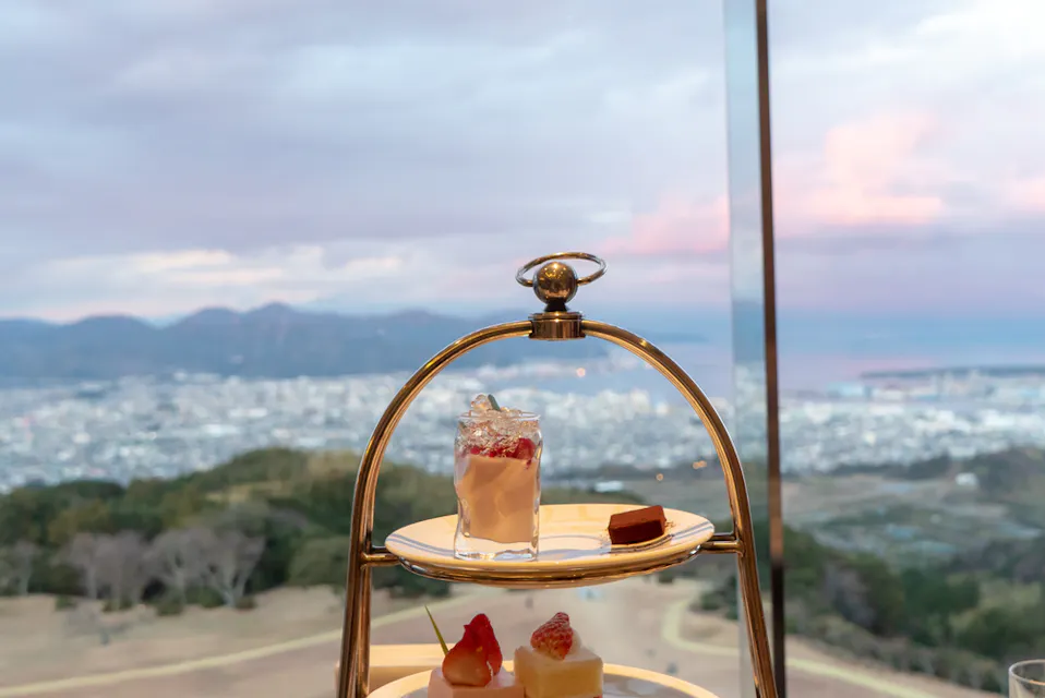 A dessert stand with various sweets: a glass with strawberries and cream, and two small cakes. It's set against a backdrop of a cityscape with hills and water under a cloudy sky seen through a large window.