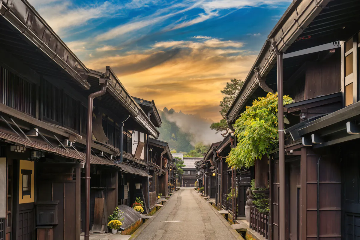 A picturesque street in a traditional Japanese village with wooden buildings lining a narrow path. The scene is framed by a colorful sunset sky and misty hills in the background, enhancing the serene atmosphere.