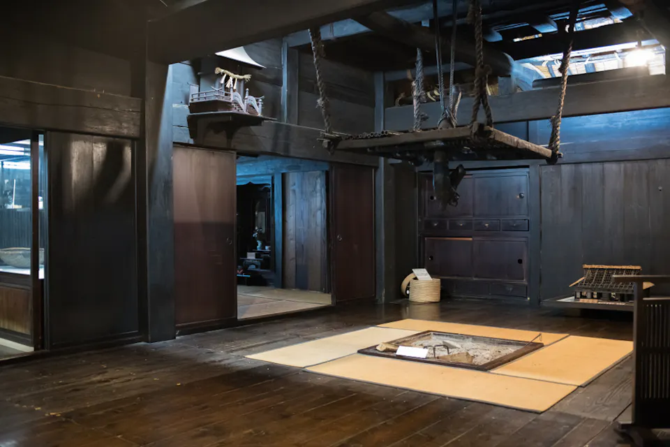 Takayama Old Town Museum A traditional Japanese room with wooden walls and floor, featuring a sunken hearth (irori) in the center, surrounded by tatami mats. A hanging kettle chain and rustic wooden furniture add to the room's historic atmosphere.