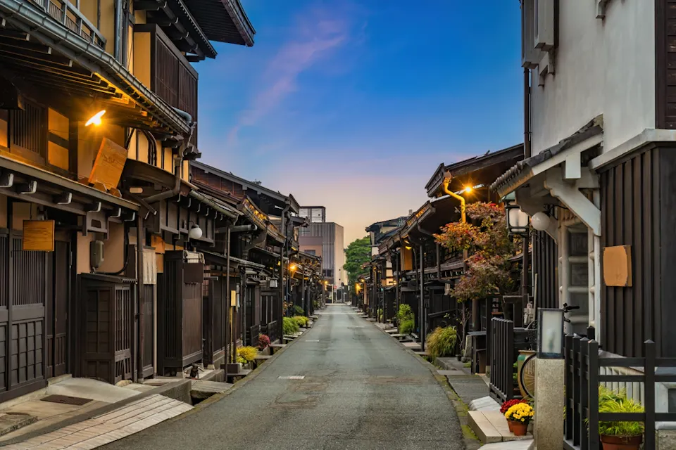 Takayama Old Town Opening Hours A narrow street lined with traditional wooden Japanese buildings, accentuated by soft evening lights. The pathway is empty, leading towards a distant sunset sky, with potted plants adding a touch of greenery to the scene.