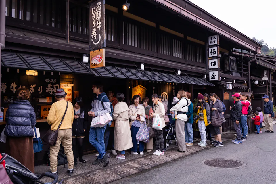 Hida Beef A group of people waiting in line outside a traditional Japanese store on a street. The shop has wooden architecture with signs in Japanese, and many in the crowd hold bags, some with children, as they queue.