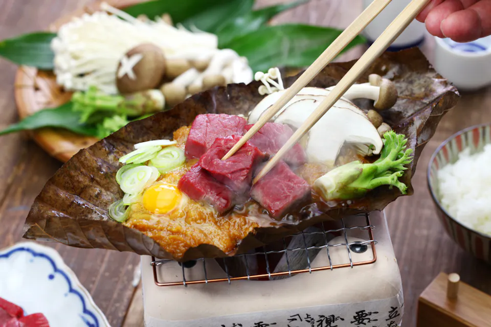 Hoba Miso A close-up of a traditional Japanese meal featuring raw beef cubes, a cracked egg, enoki mushrooms, and sliced green onions cooking on a large leaf over a small grill. A hand holds chopsticks, adjusting the beef. Sides of rice and vegetables accompany.