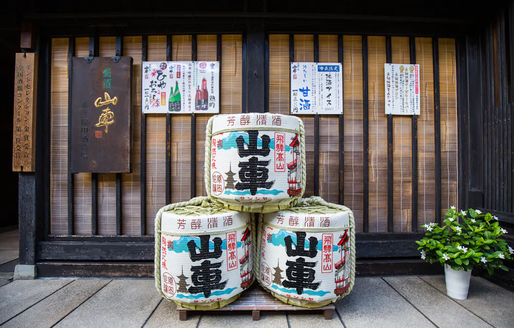 Locally Brewed Sake Three large sake barrels with Japanese characters are stacked in front of a traditional wooden structure. Above them are colorful signs with Japanese text, and a small green potted plant sits to the right.