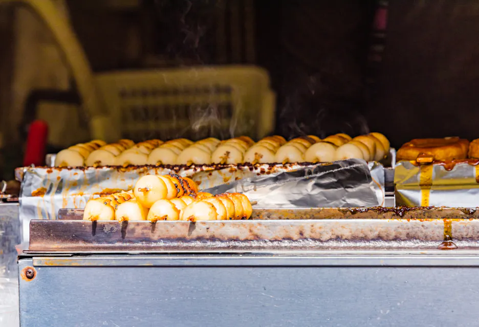 Rice Balls and Rice Crackers Rows of grilled dango (Japanese rice dumplings) on skewers, sizzling over a hot metal grill. Some skewers are brushed with sauce, creating steam that rises above the food, indicating cooking in progress.