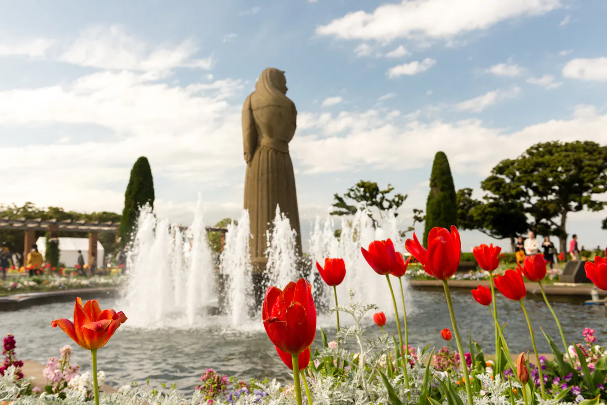 A garden scene with red tulips in the foreground, a fountain, and a large stone statue in the background. The sky is partly cloudy, and there are bushes and small trees, with people visible in the distance.