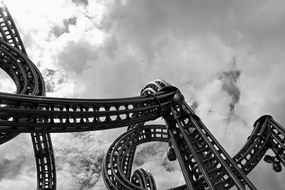 Black and white image of a complex roller coaster structure against a cloudy sky. The tracks loop and intertwine, creating an intricate design that stands out boldly against the backdrop of scattered clouds.