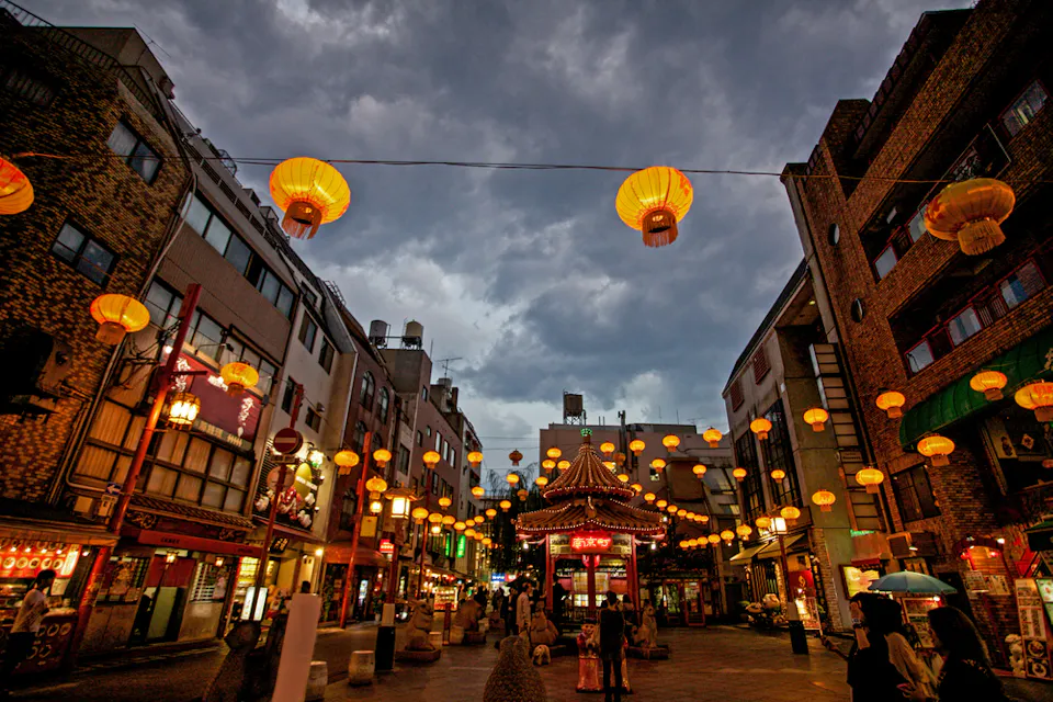 A bustling street in Chinatown at dusk, adorned with numerous glowing lanterns hung across the road. The sky is overcast, creating a dramatic backdrop for the illuminated scene. People are walking, enjoying the lively atmosphere.