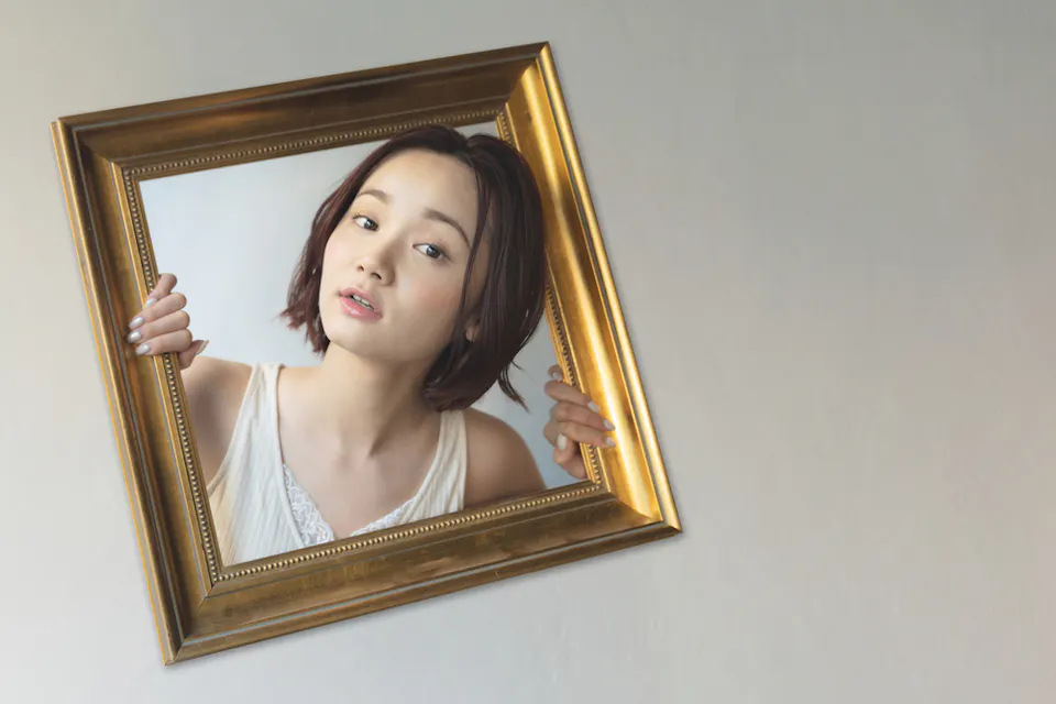 A person with short hair leans through an empty gold picture frame, creating an illusion of emerging from it. The frame is tilted against a plain background. They wear a light-colored top and have a neutral expression.