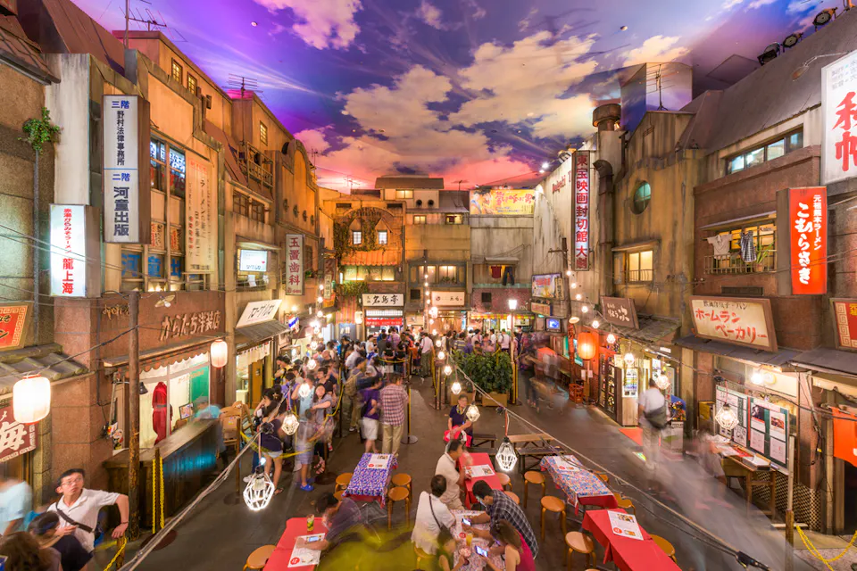A bustling indoor market mimicking a vintage Japanese street scene with vibrant lanterns, crowded tables, and people enjoying food. The ceiling is painted to resemble a sky at sunset, enhancing the lively, nostalgic atmosphere.