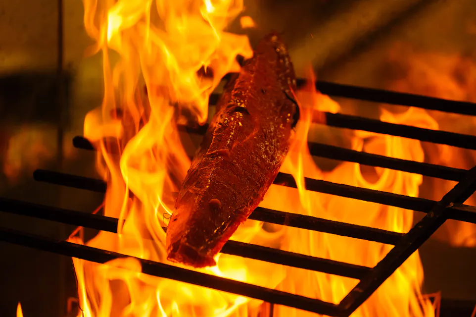 Straw Grilled Bonito A piece of salmon is being grilled over open flames. The fish is positioned on metal grates, with bright orange flames surrounding it, creating a dramatic and intense cooking scene.