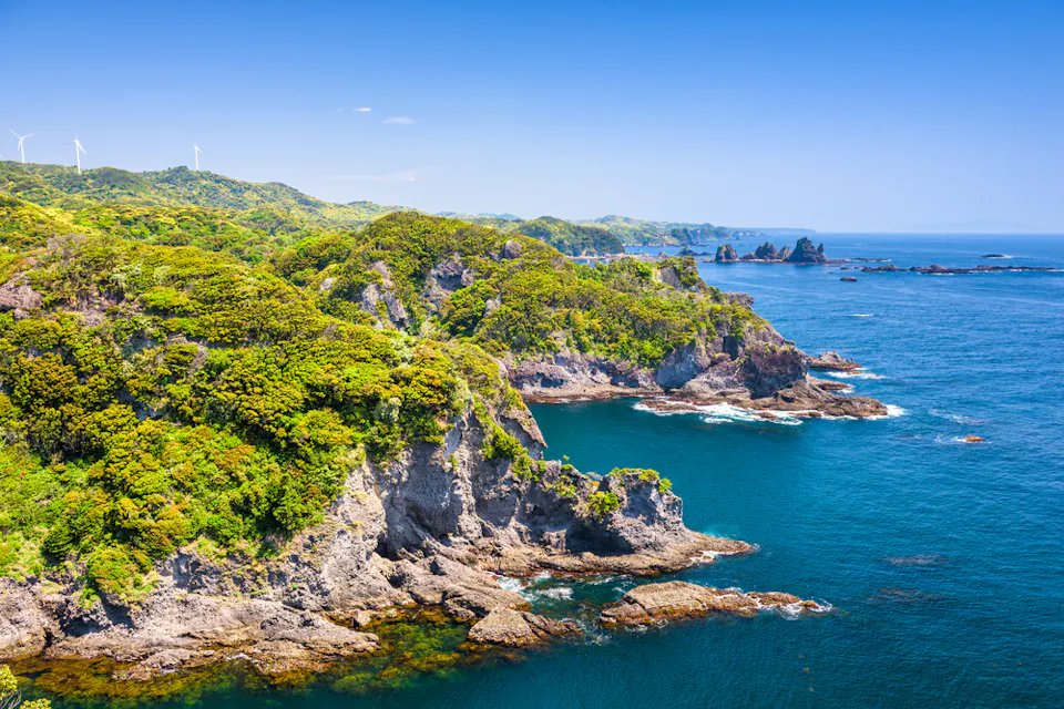 Scenic coastal landscape featuring lush green cliffs and a clear blue sea under a bright sky. Wind turbines are visible on the distant hills. Rocky formations extend into the water, creating a rugged shoreline.