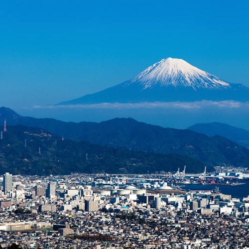 Shizuoka City A cityscape with a mix of modern buildings and industrial areas, set against a backdrop of green hills. In the distance, Mount Fuji stands majestically under a clear blue sky with a snow-capped peak.