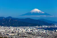 A cityscape with a mix of modern buildings and industrial areas, set against a backdrop of green hills. In the distance, Mount Fuji stands majestically under a clear blue sky with a snow-capped peak.