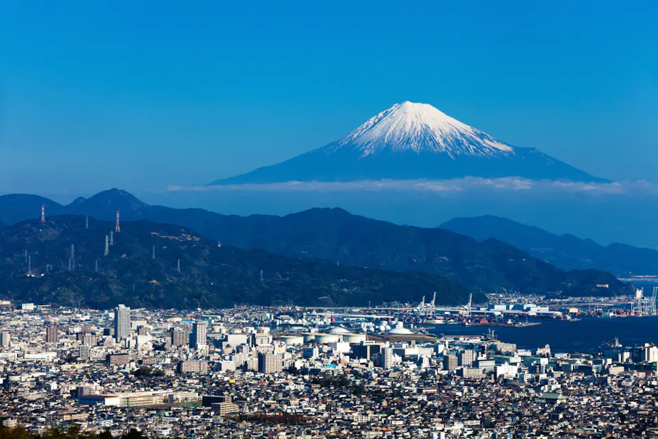 A cityscape with a mix of modern buildings and industrial areas, set against a backdrop of green hills. In the distance, Mount Fuji stands majestically under a clear blue sky with a snow-capped peak.