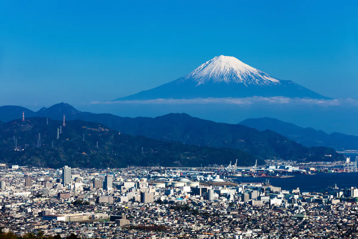 A cityscape with a mix of modern buildings and industrial areas, set against a backdrop of green hills. In the distance, Mount Fuji stands majestically under a clear blue sky with a snow-capped peak.
