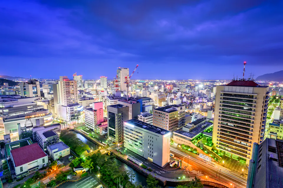 Aerial view of a vibrant cityscape at dusk with brightly lit buildings and streets, surrounded by a dark sky. Neon and street lights illuminate the scene, highlighting modern architecture and construction cranes in the distance.