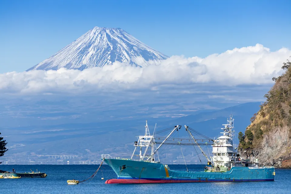 A large fishing vessel is anchored in a calm bay, with the towering, snow-capped Mount Fuji visible in the background. The sky is clear, with a few scattered clouds surrounding the mountain's peak. Nearby, a smaller boat is seen on the left.