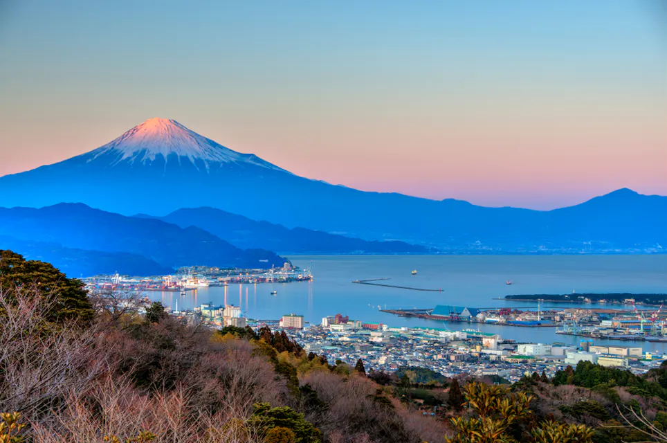 A scenic view of Mount Fuji at sunrise with a snow-capped peak. The city of Shizuoka is in the foreground, near a bay. The sky transitions from pink to blue, providing a serene and picturesque backdrop.