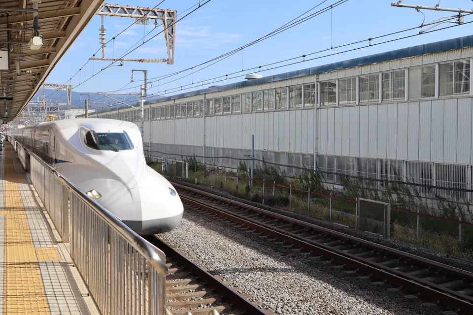 A white Shinkansen bullet train arrives at a station in Japan on a clear day. The platform is empty, and train tracks run parallel to a large industrial building. Overhead power lines stretch across the scene.