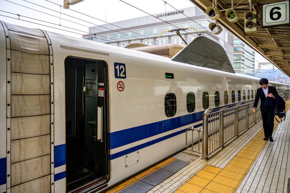 A man in a suit walks beside a white and blue high-speed train at a station. The train is on platform 6, and its doors are open. Overhead awnings and station infrastructure are visible.