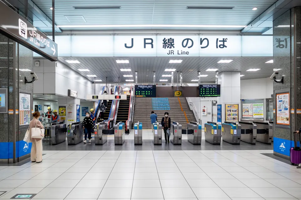 A modern subway station entrance for JR Line with turnstiles and digital display boards. People are walking through the area, and the space is brightly lit with blue and grey accents. An escalator and stairs are in the background.