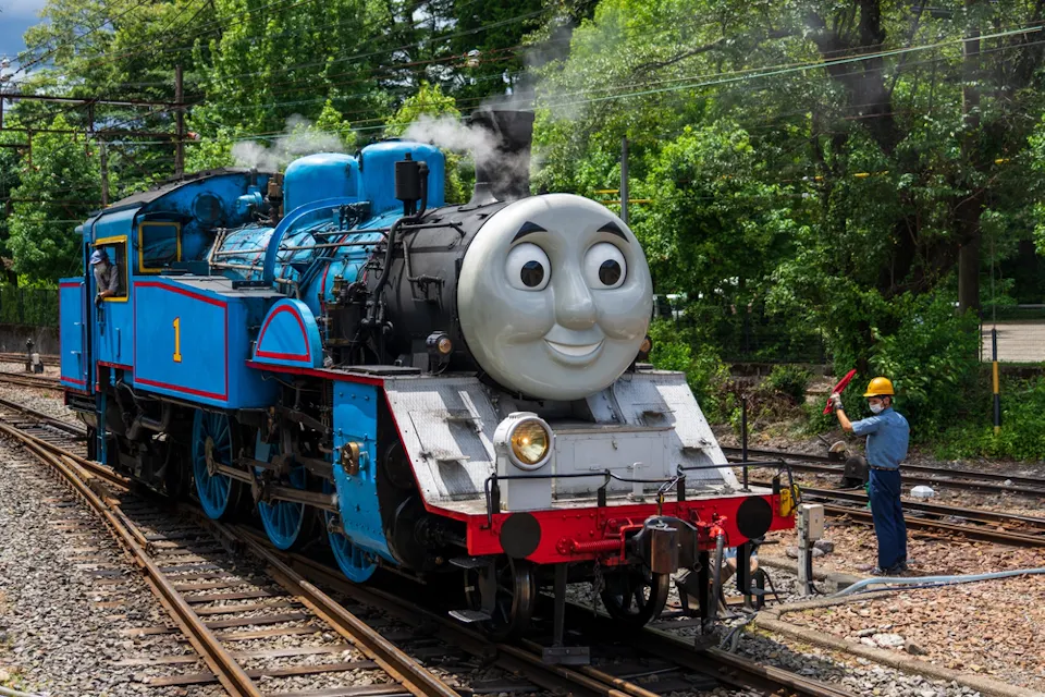 A blue steam locomotive with a smiling face on the front is on railway tracks. A worker in a hard hat stands nearby, holding a signal paddle. Green trees and power lines are in the background.